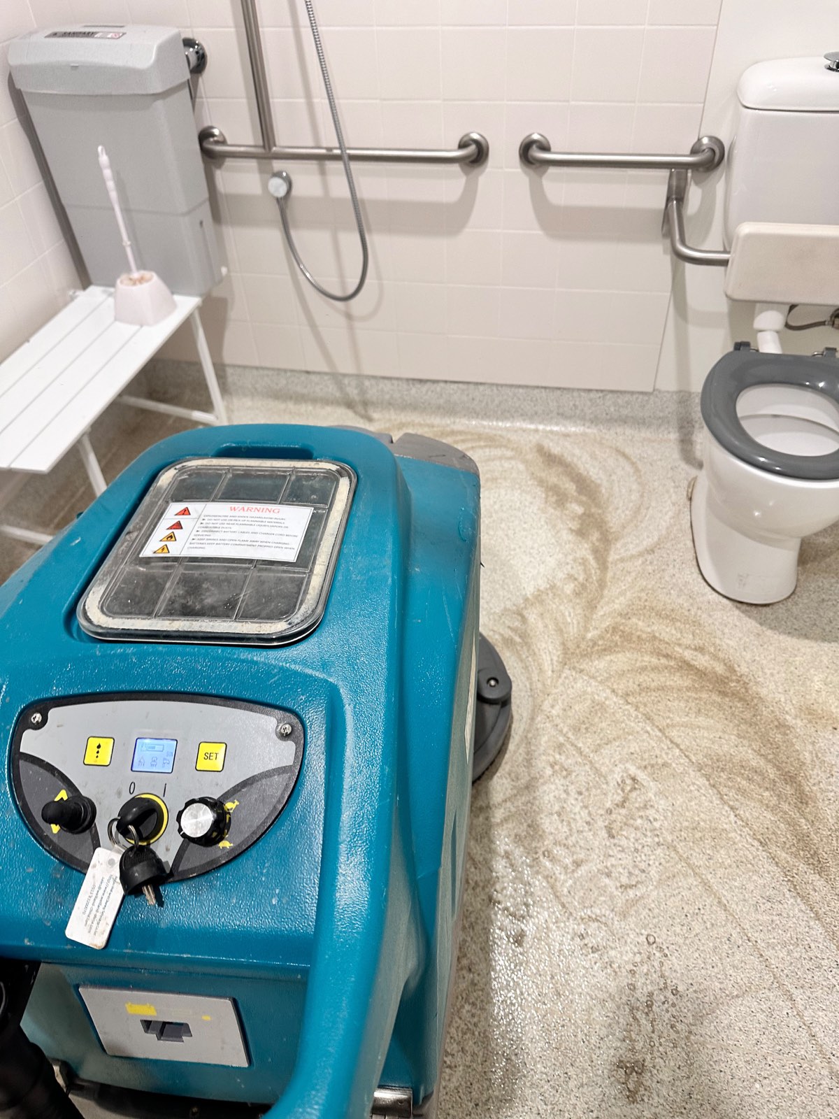 Accessible bathroom with grab rails being scrubbed by a floor machine